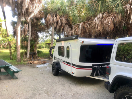 Your rv rental adventure awaits! A white travel trailer hitched to a Jeep at a palm tree campsite with a picnic table.