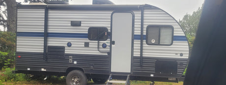 Side view of a gray and blue travel trailer, an RV rental unit. It has a prominent door, windows, and outdoor speakers.