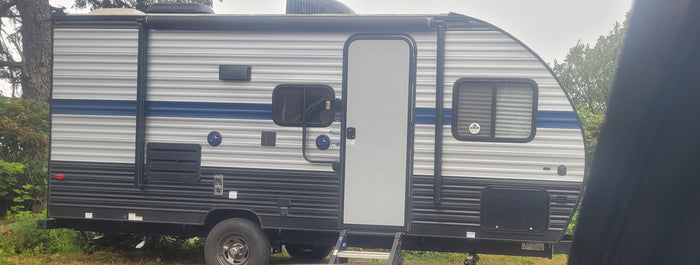 Side view of a gray and blue travel trailer, an RV rental unit. It has a prominent door, windows, and outdoor speakers.