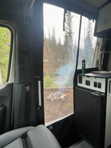 RV rental camp setup: A view from inside a camper van shows a smoking campfire in a forest, with a fridge and sink visible.