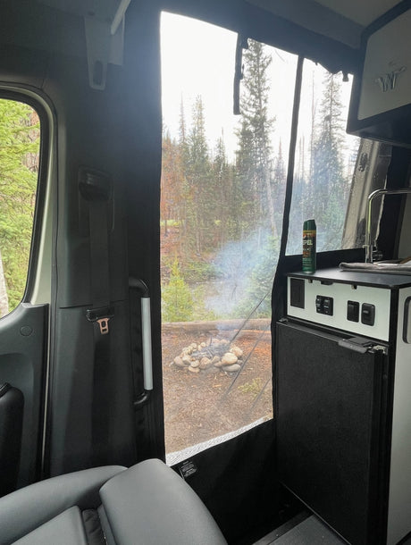 RV rental camp setup: A view from inside a camper van shows a smoking campfire in a forest, with a fridge and sink visible.