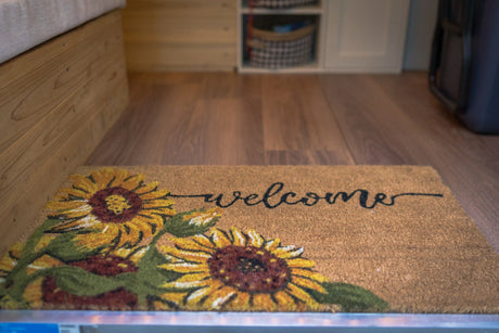 A 'welcome' mat with sunflowers adds a cozy touch to an RV rental interior, featuring wood floors and built-in storage.