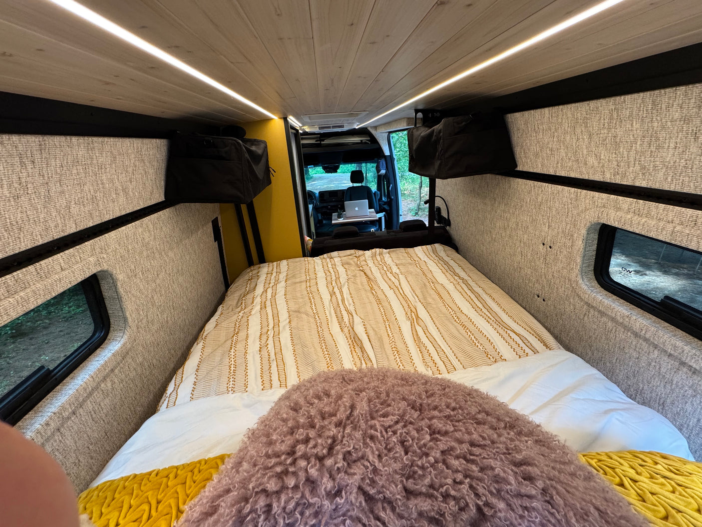 Cozy RV rental interior: Bed, desk w/ laptop, and bright lighting for comfortable van life travel.