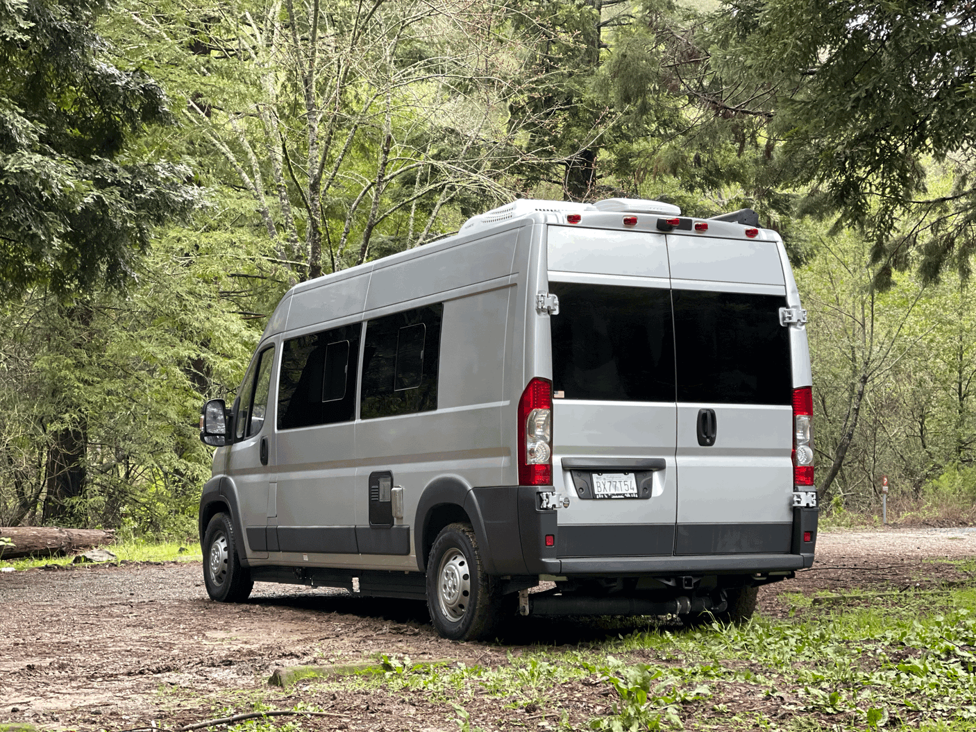 Silver RV rental camper van parked on a dirt path amidst lush forest. Ideal for nature and outdoor adventures.