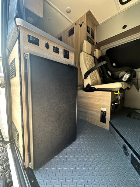 RV rental interior: black refrigerator, beige patterned seat with child car seat, controls on wood-grain cabinets, textured floor.