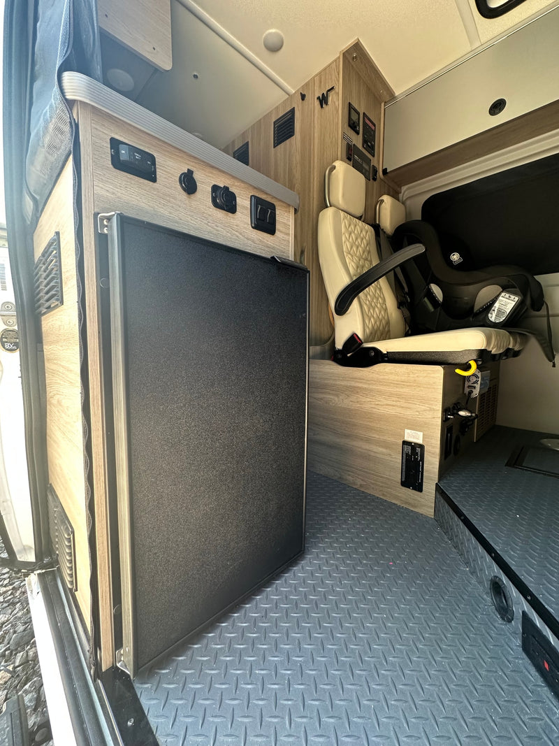 RV rental interior: black refrigerator, beige patterned seat with child car seat, controls on wood-grain cabinets, textured floor. — photo 8