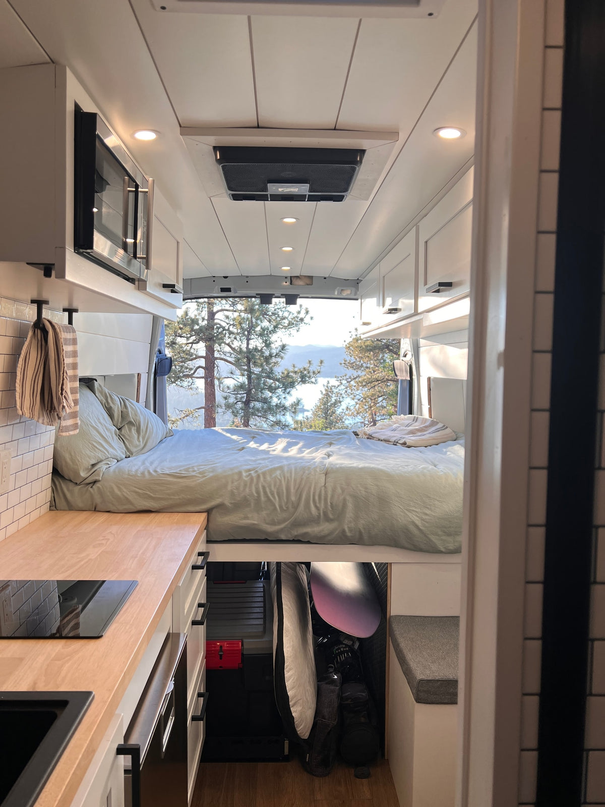 Cozy rv rental interior: van bed overlooks a scenic lake & pine trees, with a compact kitchen in foreground. — photo 8