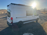 White RV rental camper van parked on gravel, strong sunflare, roof rack, hills, and building in background at sunset.