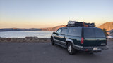 This dark green Chevrolet Suburban, outfitted like an RV rental, is parked overlooking a vast lake and mountains at sunrise.