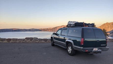 This dark green Chevrolet Suburban, outfitted like an RV rental, is parked overlooking a vast lake and mountains at sunrise.