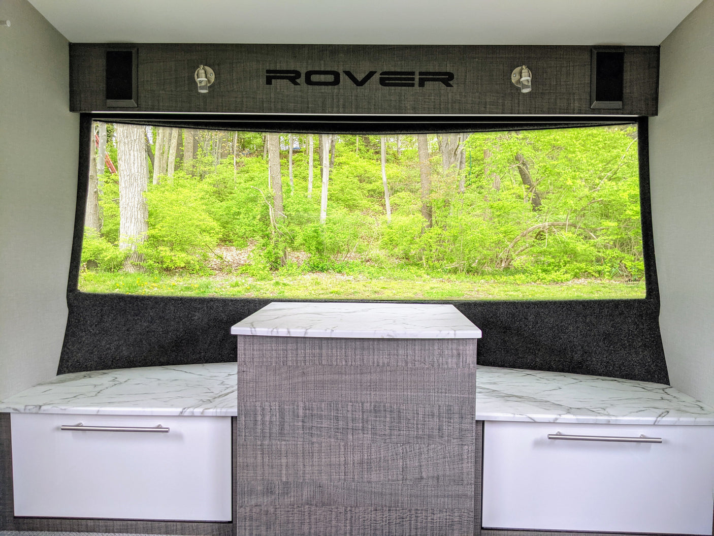 Bright RV rental interior with forest view. Features built-in bench seating, a central table, and "ROVER" branding.