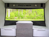 Bright RV rental interior with forest view. Features built-in bench seating, a central table, and "ROVER" branding.