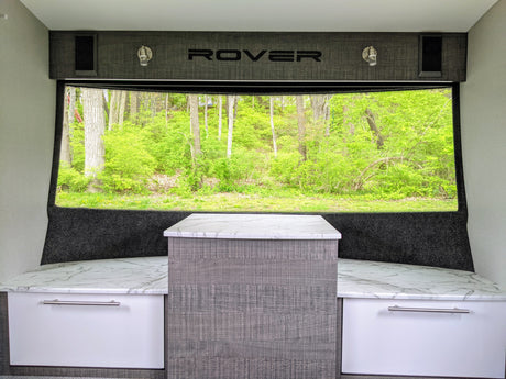 Bright RV rental interior with forest view. Features built-in bench seating, a central table, and "ROVER" branding.