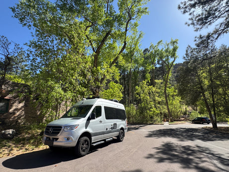Silver Mercedes Sprinter rv rental camper van parked on an asphalt road, surrounded by vibrant green trees under a clear blue sky.