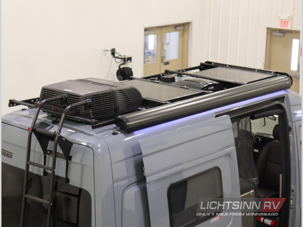 A modern gray camper van for RV rental, featuring rooftop AC, solar panels, ladder, and a blue-lit awning.