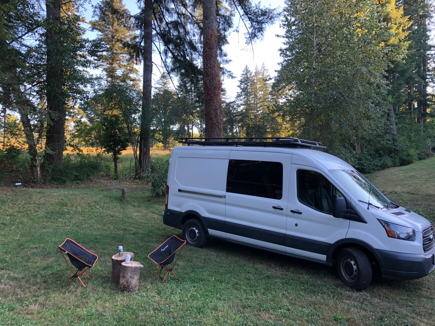 White Ford Transit camper van (RV rental) on grass with camp chairs and tree stump tables, set in a wooded area. — photo 6