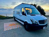 White RV rental van by the ocean at sunset with a sun mat on the sand. Perfect for coastal adventures.