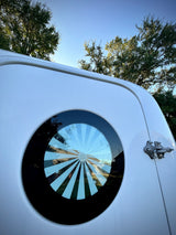 A white RV features a unique round window with a sunburst pattern, reflecting trees and blue sky. Ideal for your next rv rental.
