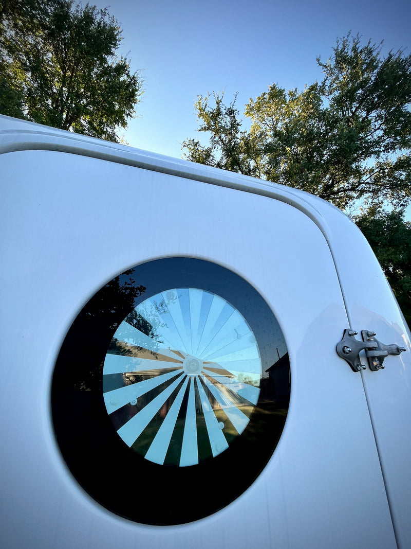 A white RV features a unique round window with a sunburst pattern, reflecting trees and blue sky. Ideal for your next rv rental. — photo 11