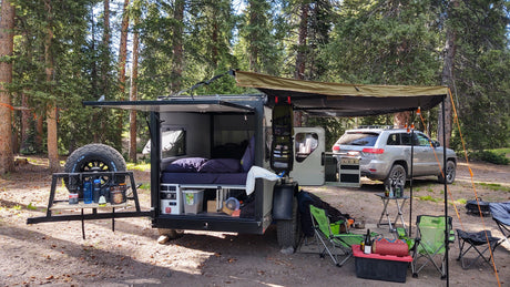 RV rental adventure: Off-road camper trailer & SUV parked in a forest, fully set up for cozy camping with an outdoor kitchen.