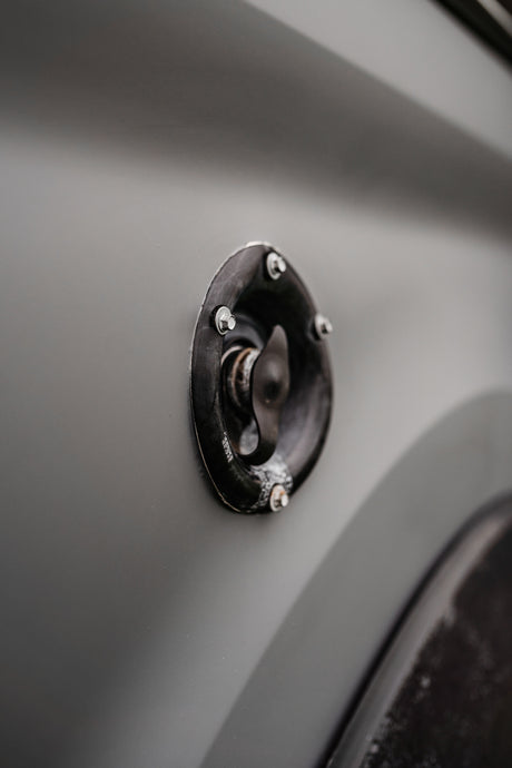 Black access cap on a grey RV rental's side, likely for fuel or water. Functional vehicle detail.