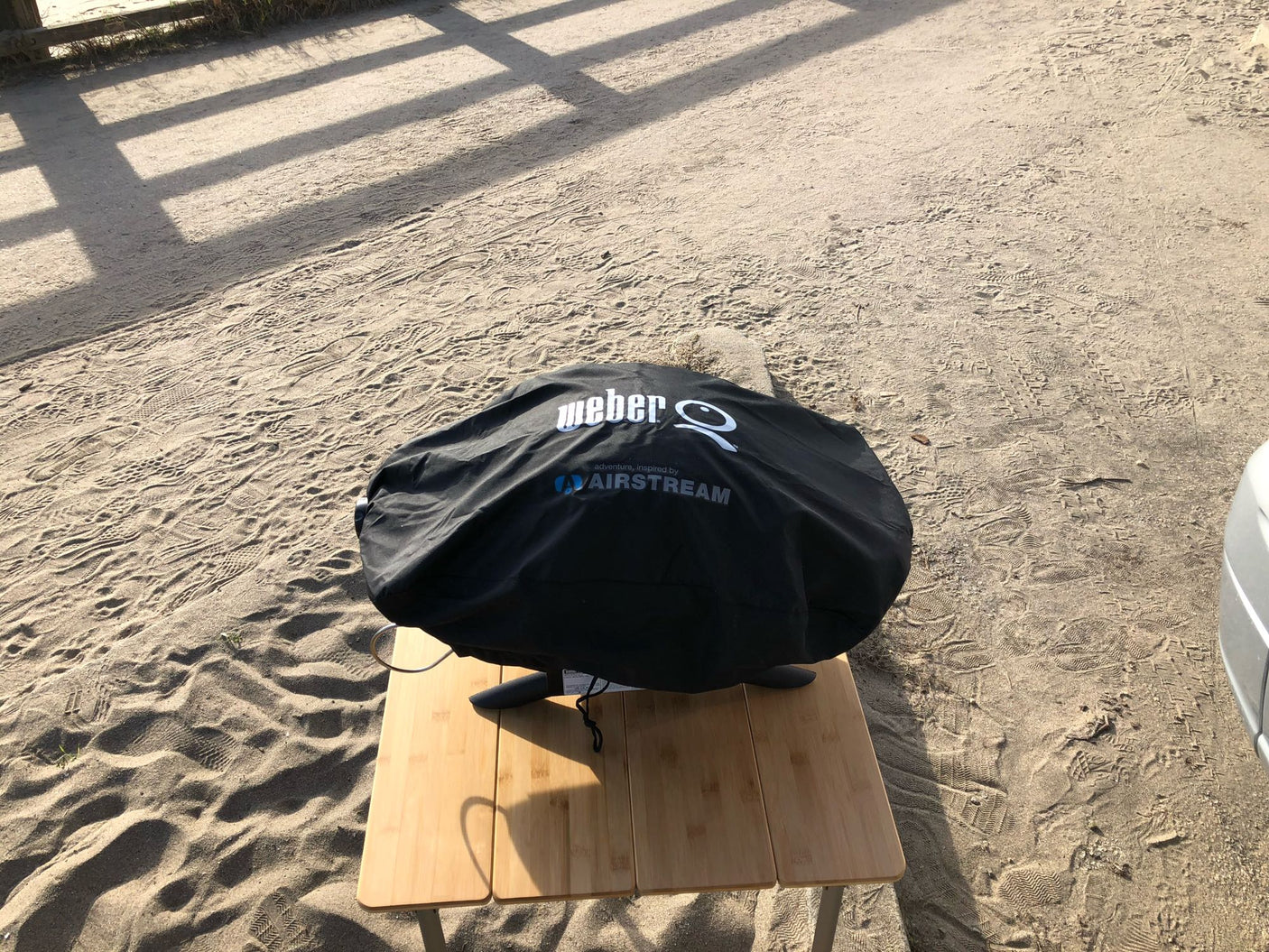 A covered Weber Airstream grill sits on a bamboo table at a sandy RV rental campsite, ready for outdoor meals.