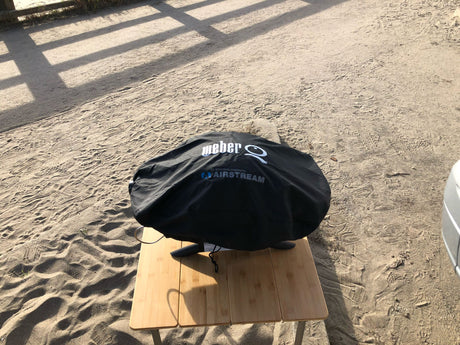 A covered Weber Airstream grill sits on a bamboo table at a sandy RV rental campsite, ready for outdoor meals.