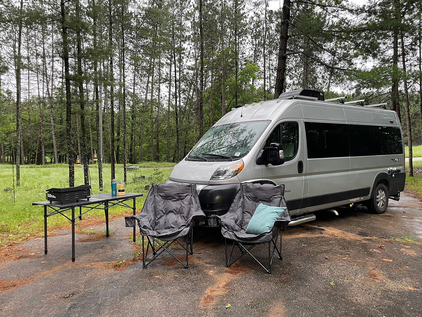 RV rental parked in a damp forest clearing, featuring a camper van, two chairs, and a table with a grill.