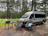 RV rental parked in a damp forest clearing, featuring a camper van, two chairs, and a table with a grill.
