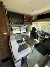 Inside an RV rental: view of the driver's area, dining booth with table, and overhead entertainment system.
