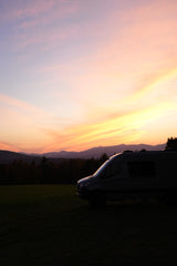 Gorgeous sunset behind an RV rental, parked on a field with silhouetted mountains. Sky glows with pink, orange, and yellow clouds.