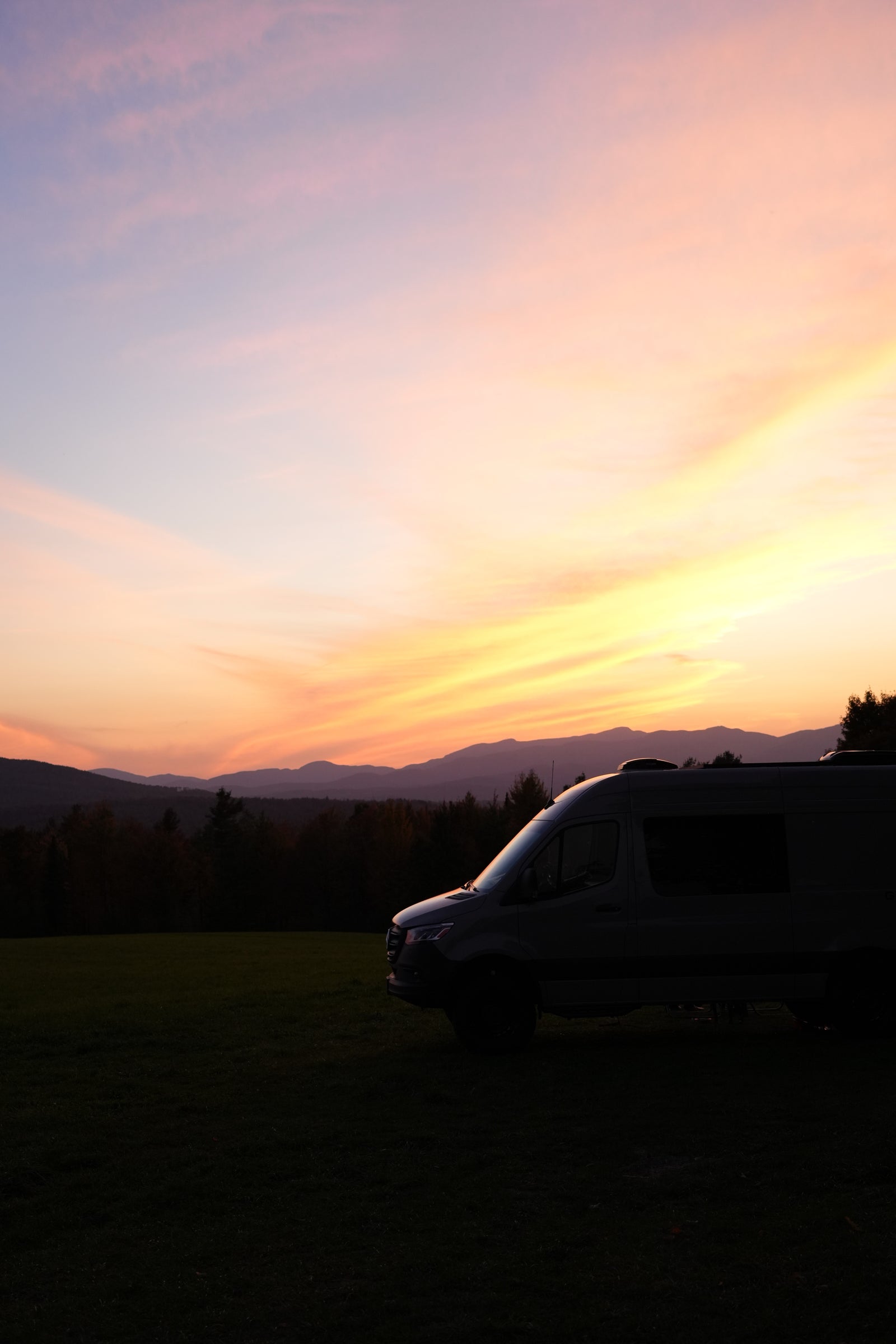Gorgeous sunset behind an RV rental, parked on a field with silhouetted mountains. Sky glows with pink, orange, and yellow clouds.