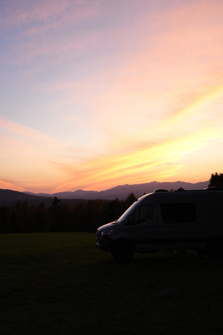 Gorgeous sunset behind an RV rental, parked on a field with silhouetted mountains. Sky glows with pink, orange, and yellow clouds.