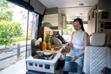 Cozy rv rental interior: woman pours coffee in the compact kitchen with stove, sink, and fruit. Ideal for adventure travel.