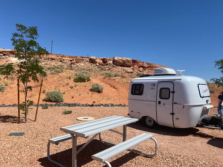 A white Scamp travel trailer (RV rental) at a sunny desert campsite with a picnic table.