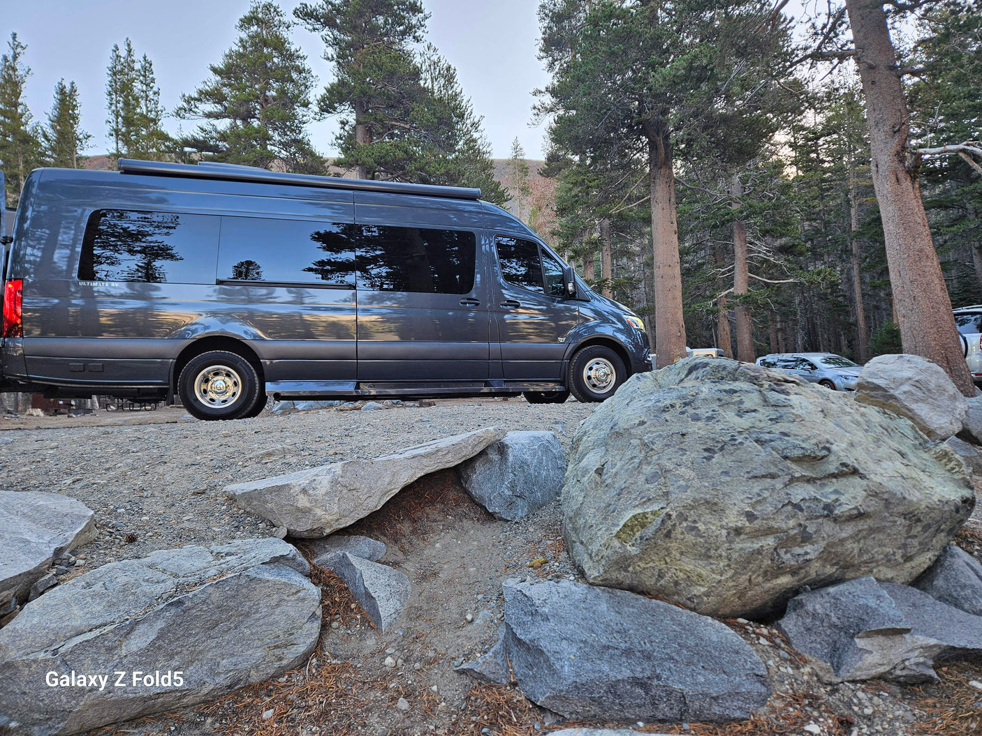 An Ultimate RV camper van, a perfect rv rental, parked on gravel amidst large rocks and pine trees.