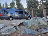 An Ultimate RV camper van, a perfect rv rental, parked on gravel amidst large rocks and pine trees.