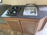 This RV rental features a compact kitchen with a two-burner stovetop, sink containing new manuals/keys, and wooden cabinets.