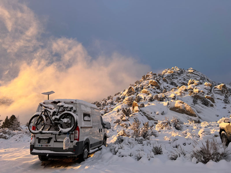 A white rv rental van with bikes and a Starlink dish, covered in snow, parked in a golden-lit winter mountain scene. — photo 3