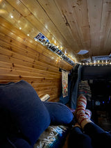 Cozy RV rental interior with warm wood panels, string lights, and photos. Person relaxing, enjoying van life comfort.