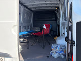 RV rental cargo van interior: cot, red chair, water bottles, and storage box on a carpeted floor.