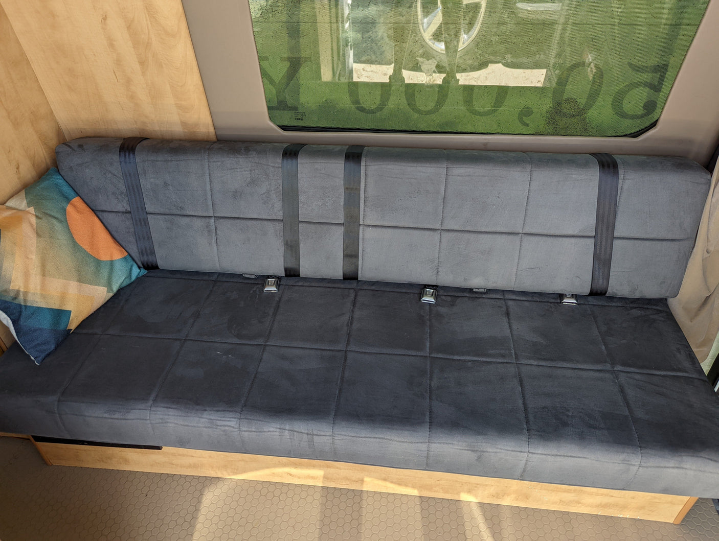 RV rental interior shows a gray bench seat with black seatbelts, a patterned pillow, and wooden walls next to a window.
