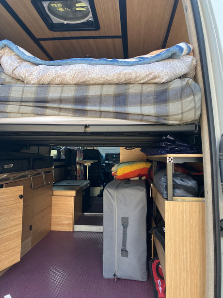 An rv rental camper van's interior, showing a raised bed, wooden cabinets, organized storage, and a roof vent.