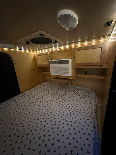 Comfortable RV rental interior featuring a bed with turtle-patterned sheets, AC unit, and ambient string lights.