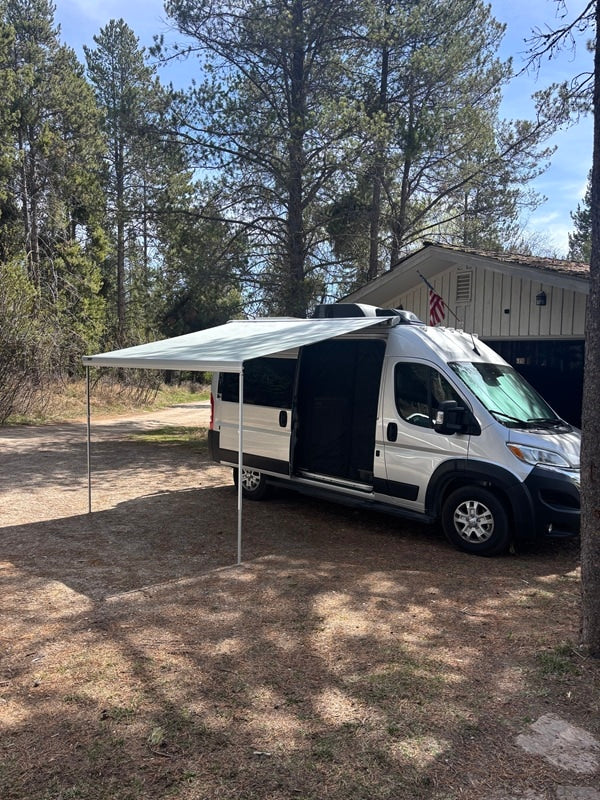 This silver RV rental, with its extended awning, is parked in a wooded setting next to a house, perfect for outdoor adventures.