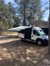 This silver RV rental, with its extended awning, is parked in a wooded setting next to a house, perfect for outdoor adventures.