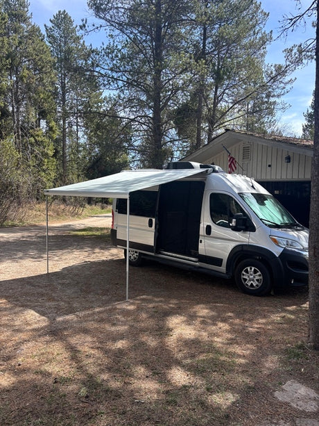 This silver RV rental, with its extended awning, is parked in a wooded setting next to a house, perfect for outdoor adventures.
