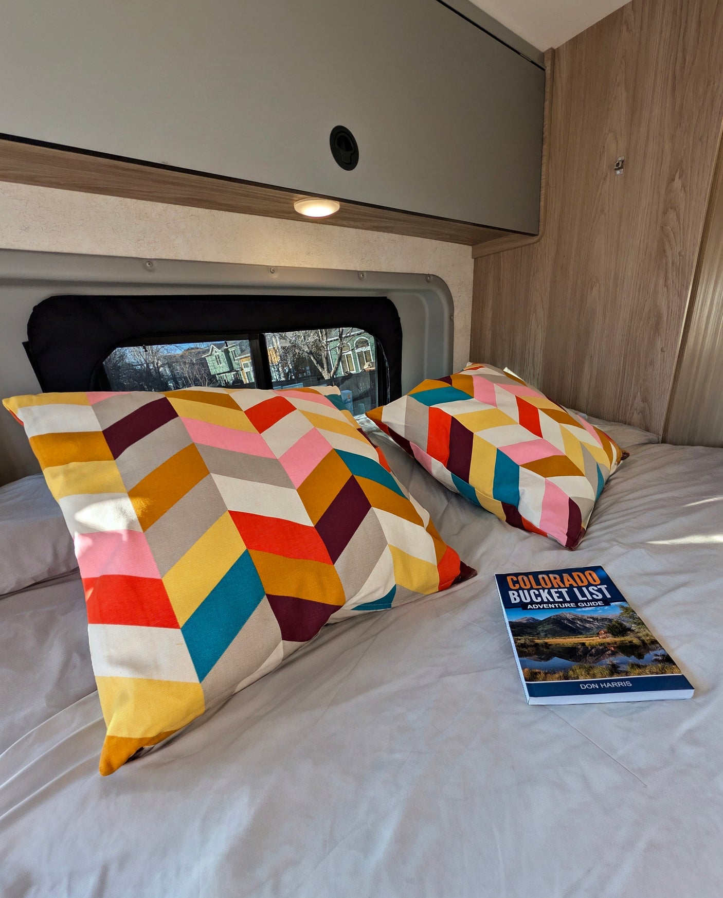 Cozy RV rental bed with vibrant geometric pillows & a 'Colorado Bucket List' guide. Ready for your next adventure!