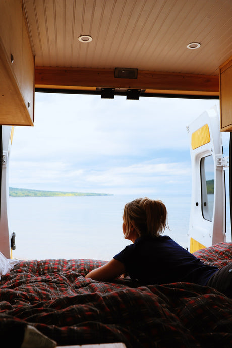 Campervan interior: A woman lies on a plaid bed, enjoying the peaceful lake view from her RV rental's open doors.