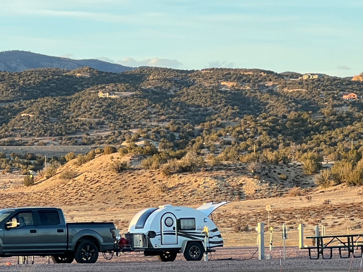 An RV rental: a white teardrop trailer is hitched to a gray pickup truck at a desert campsite, with mountains in the sunny background.
